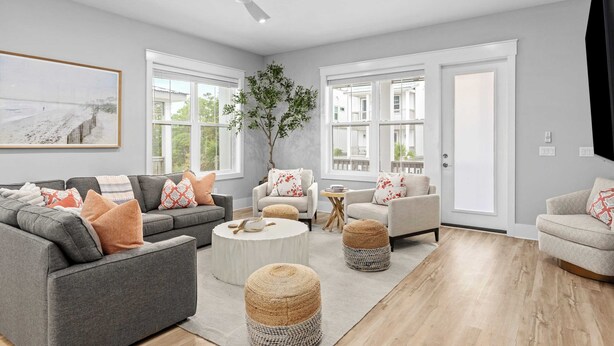 Ein modernes Wohnzimmer mit einem grauen Ecksofa, zwei Sesseln, einem runden Couchtisch und Poufs. Ein großes Fenster bietet Blick auf ein Gebäude und einen Baum.