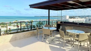 Un balcón con sillas de mimbre y una mesa de vidrio con vistas a la playa y al paisaje urbano.