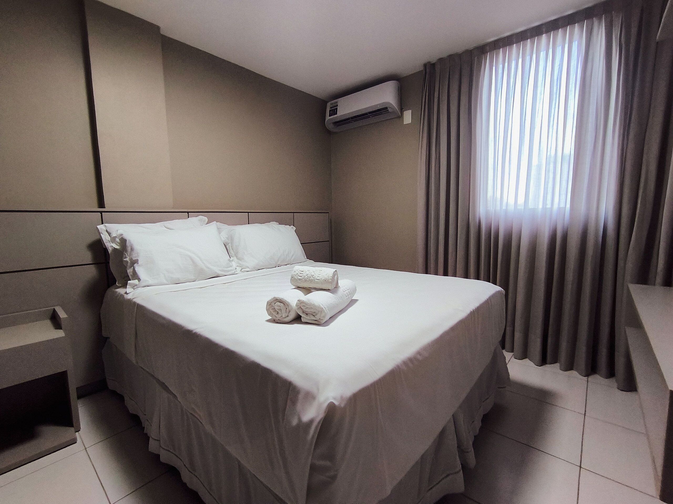 A neatly made bed with white linens and towels, a wall-mounted air conditioner, and a window with curtains.