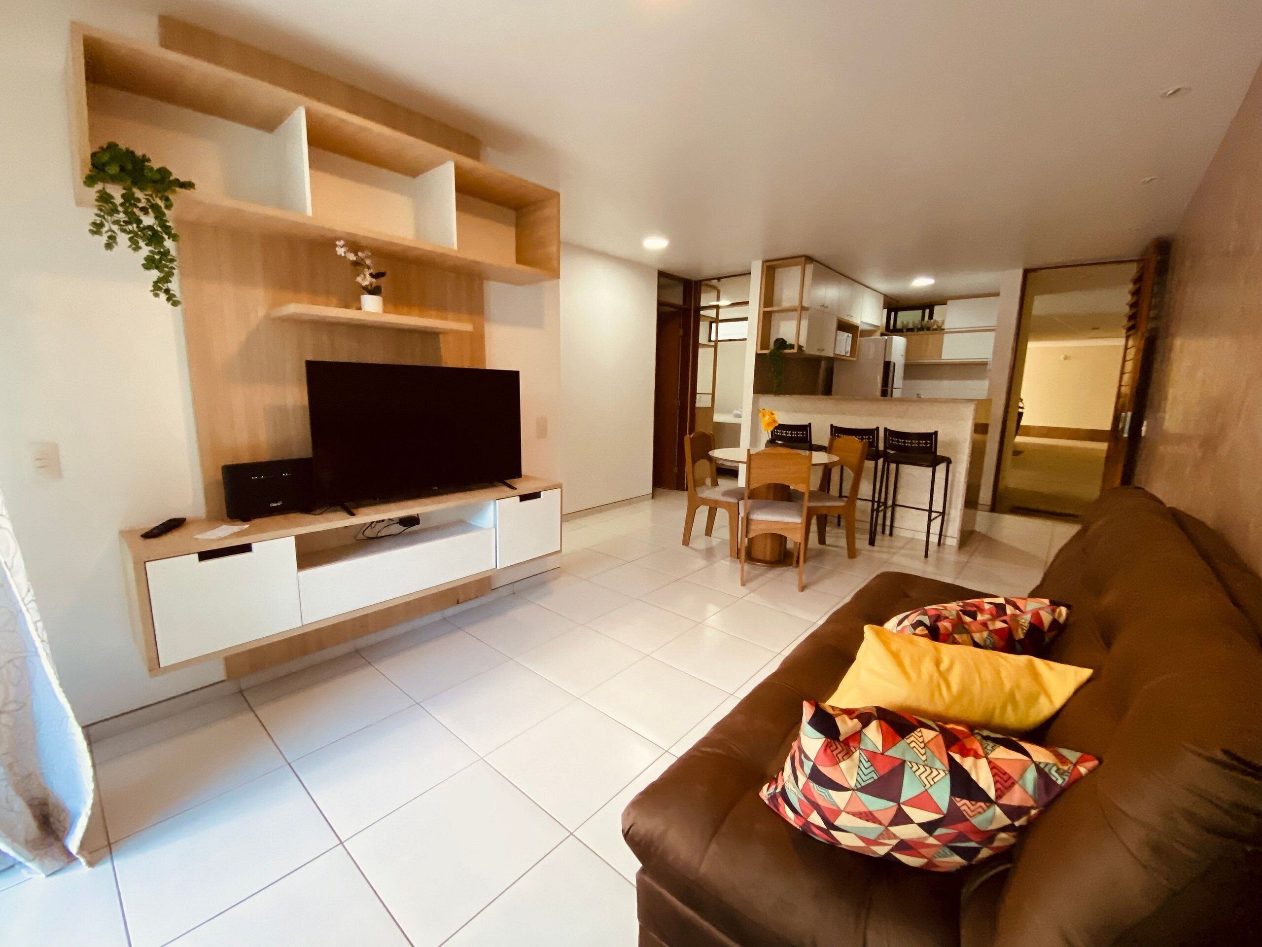 Un salon moderne avec un téléviseur à écran plat posé sur un meuble bas en bois, un canapé en cuir marron agrémenté de coussins à motifs, et un coin repas avec des chaises en bois et une petite table.