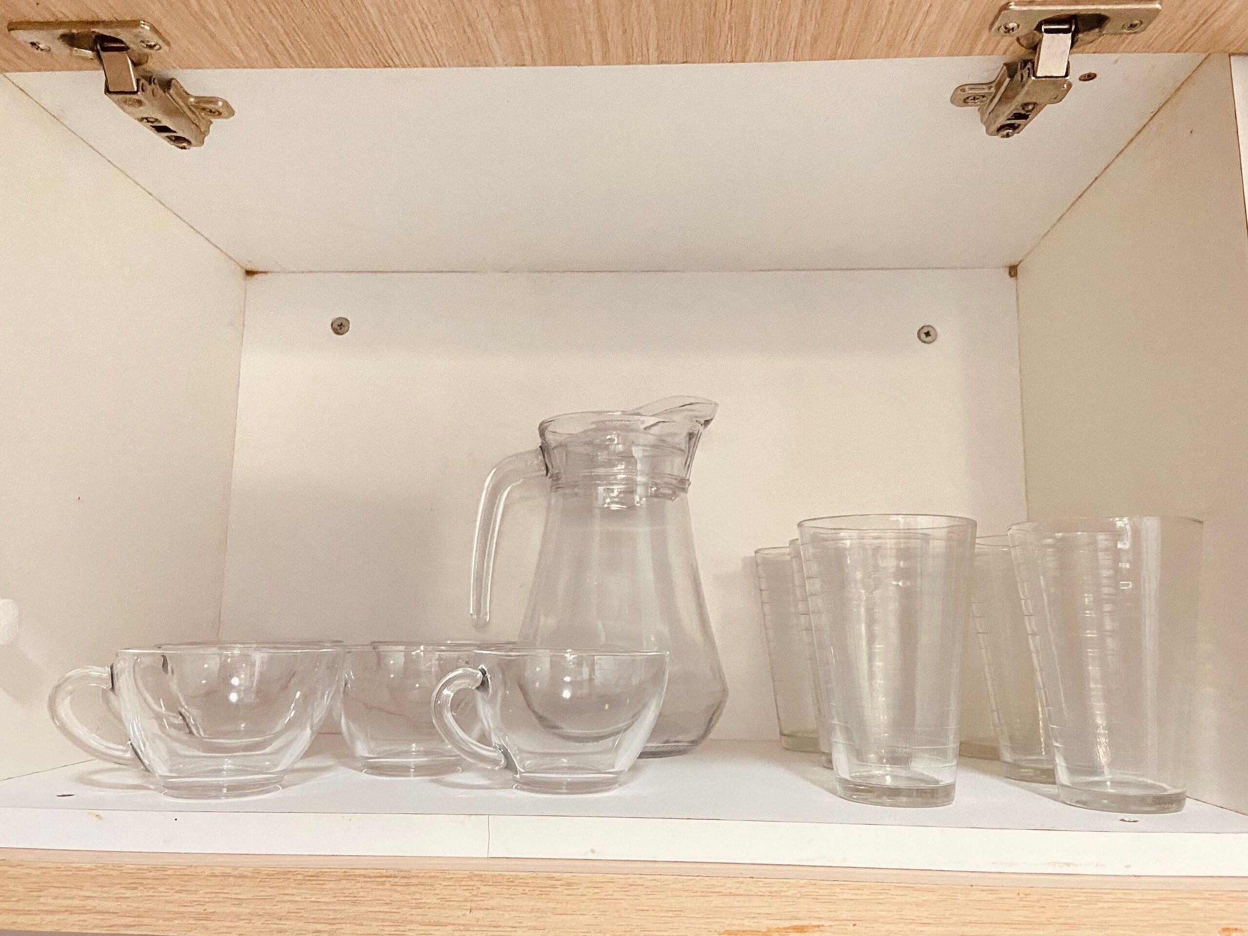 En hylla med en genomskinlig glasbringa, flera genomskinliga glas och en genomskinlig glasvas.