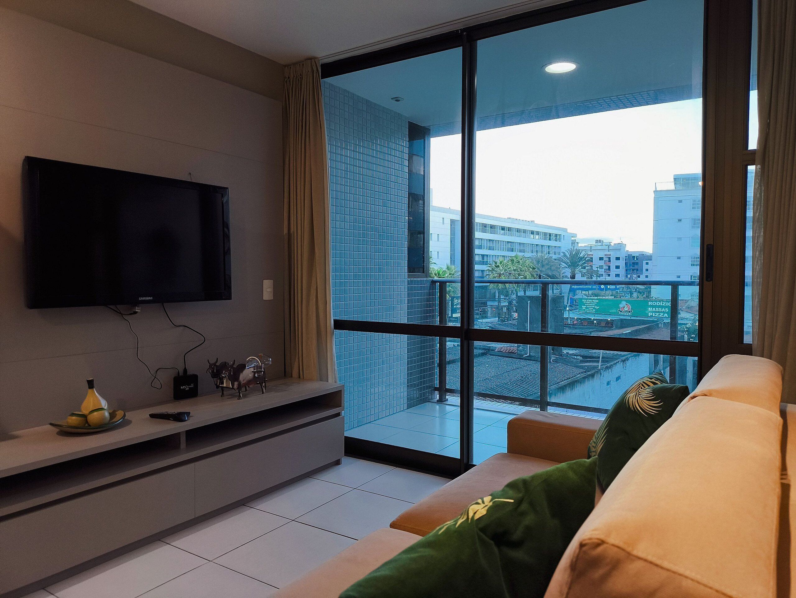 Una sala de estar moderna con un televisor de pantalla plana, un sofá con cojines verde y beige, y un ventanal con vistas a la ciudad.