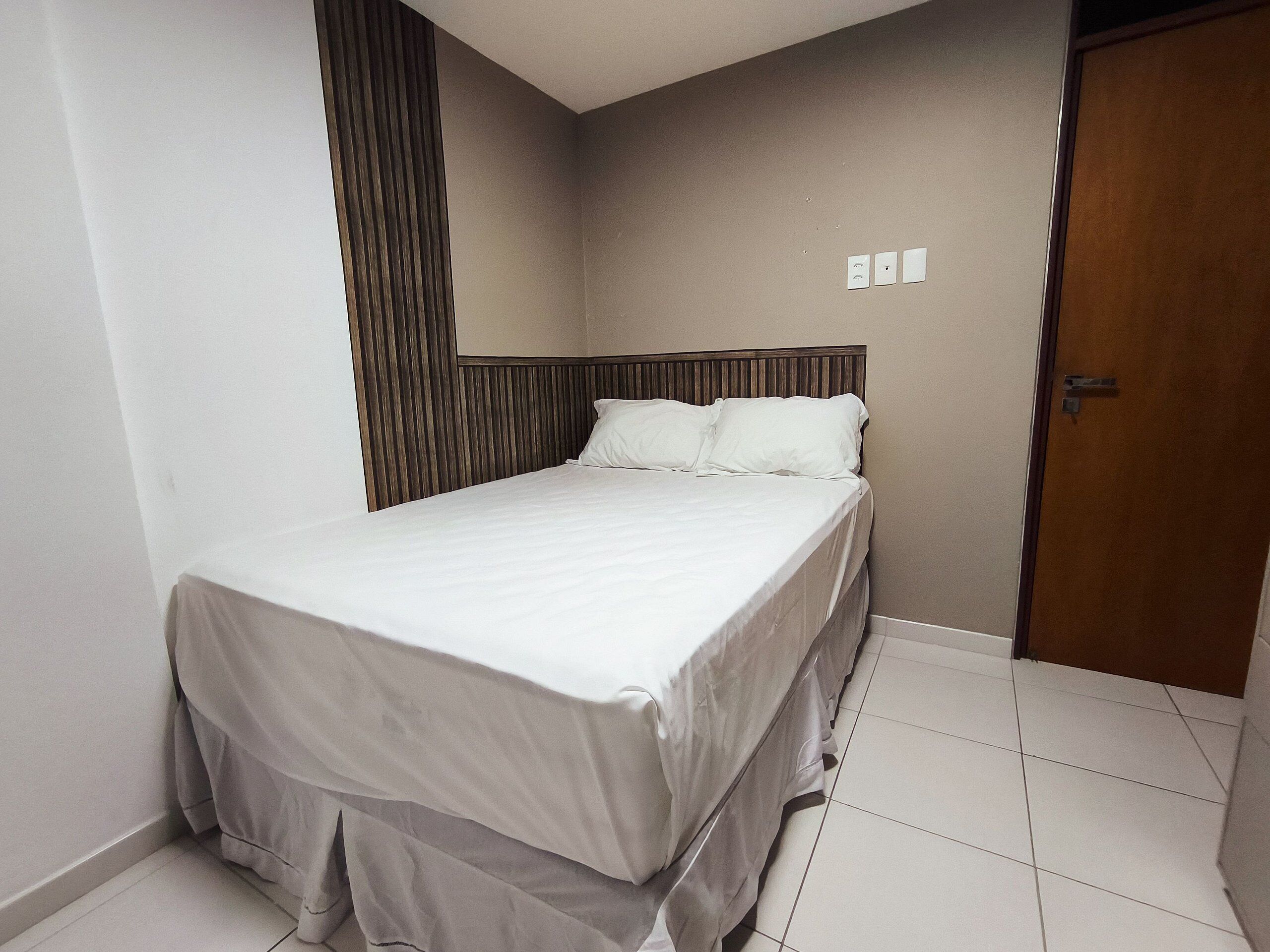 A neatly made bed in a hotel room with a wooden headboard, a door, and a tiled floor.