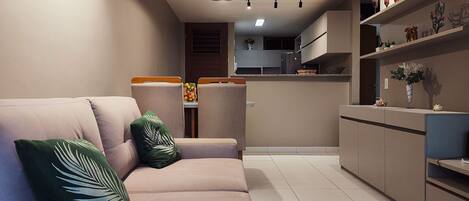 A modern living room with a sofa, a small table with a bowl of fruit, and a kitchen in the background.