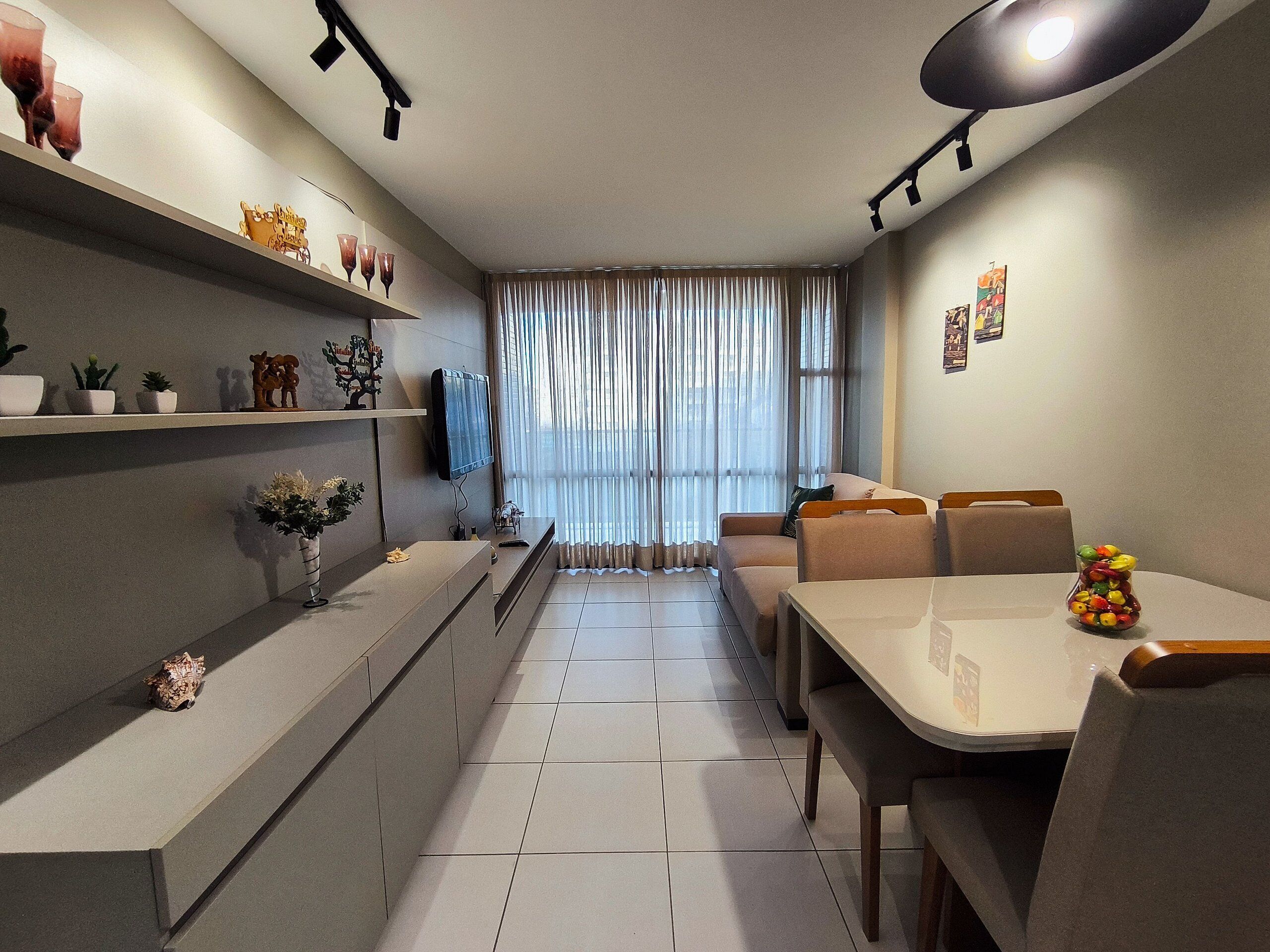 Un salon moderne avec un canapé, une table à manger et un téléviseur. On y trouve des étagères ornées d’objets décoratifs et une fenêtre avec des rideaux.