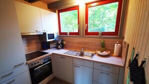 Une cuisine compacte avec des meubles blancs, une cuisinière, un four à micro-ondes et un évier. Il y a deux fenêtres avec des cadres rouges, et une petite plante sur le rebord de la fenêtre.