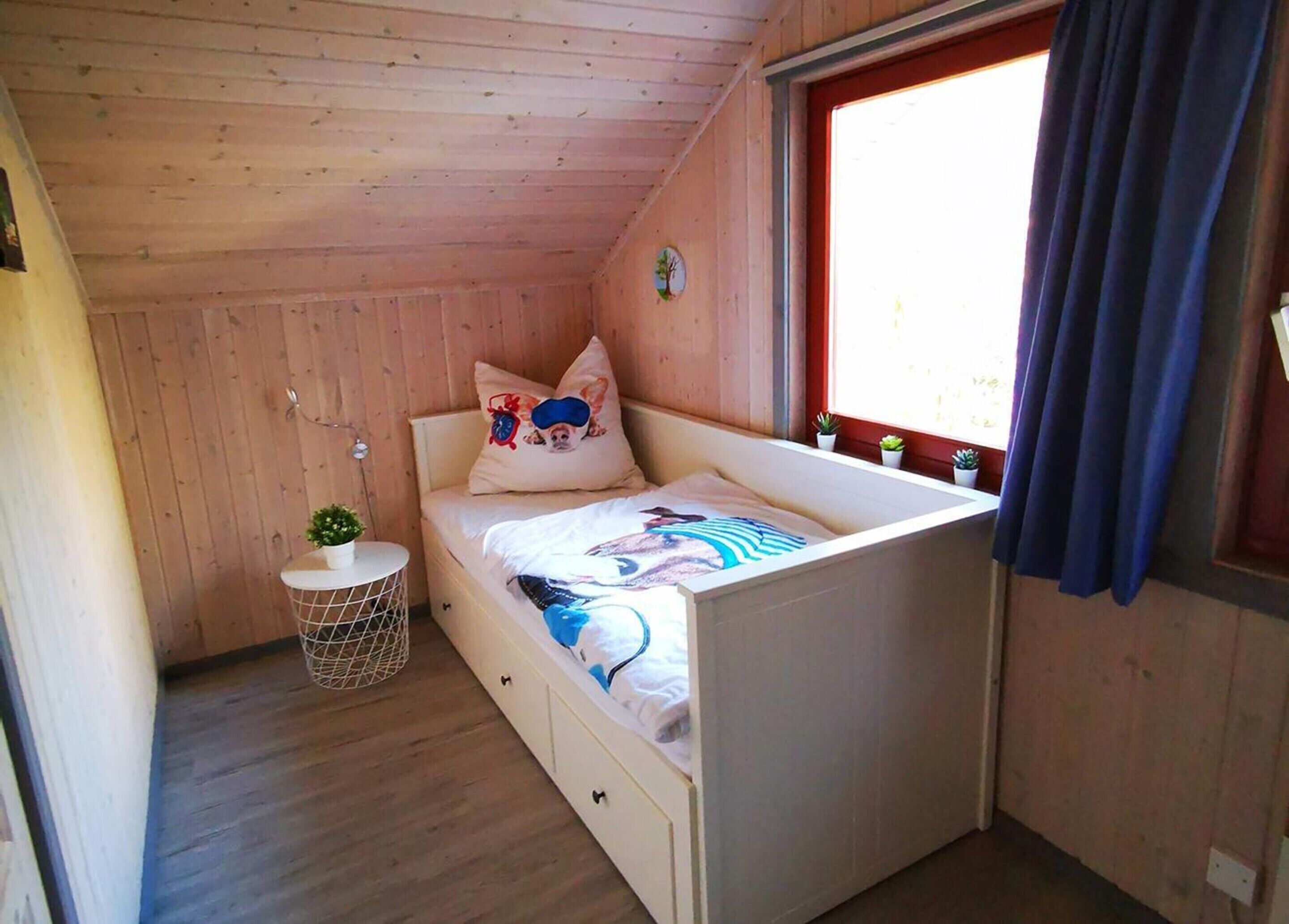 A small, cozy room with a wooden ceiling and walls, a single bed with a colorful bedspread, a small round table with a plant, and a window with curtains.