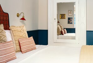 Un dormitorio con cama, almohadas, una lámpara roja en la pared, una puerta blanca y un espejo que refleja otra habitación.