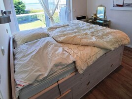 A bed with a white mattress and beige bedding, a small bedside table with a lamp, and a window with curtains.