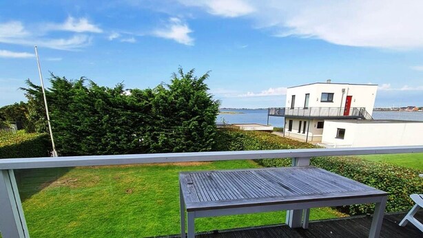 A balcony with a table and chairs, overlooking a body of water and a white building with a red door. - Haus am Merr auf Fehmarn OT Westerbergen (Fehmarn)