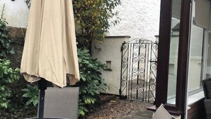 A glass table with a metal frame, a beige umbrella, and a metal gate in a garden setting.