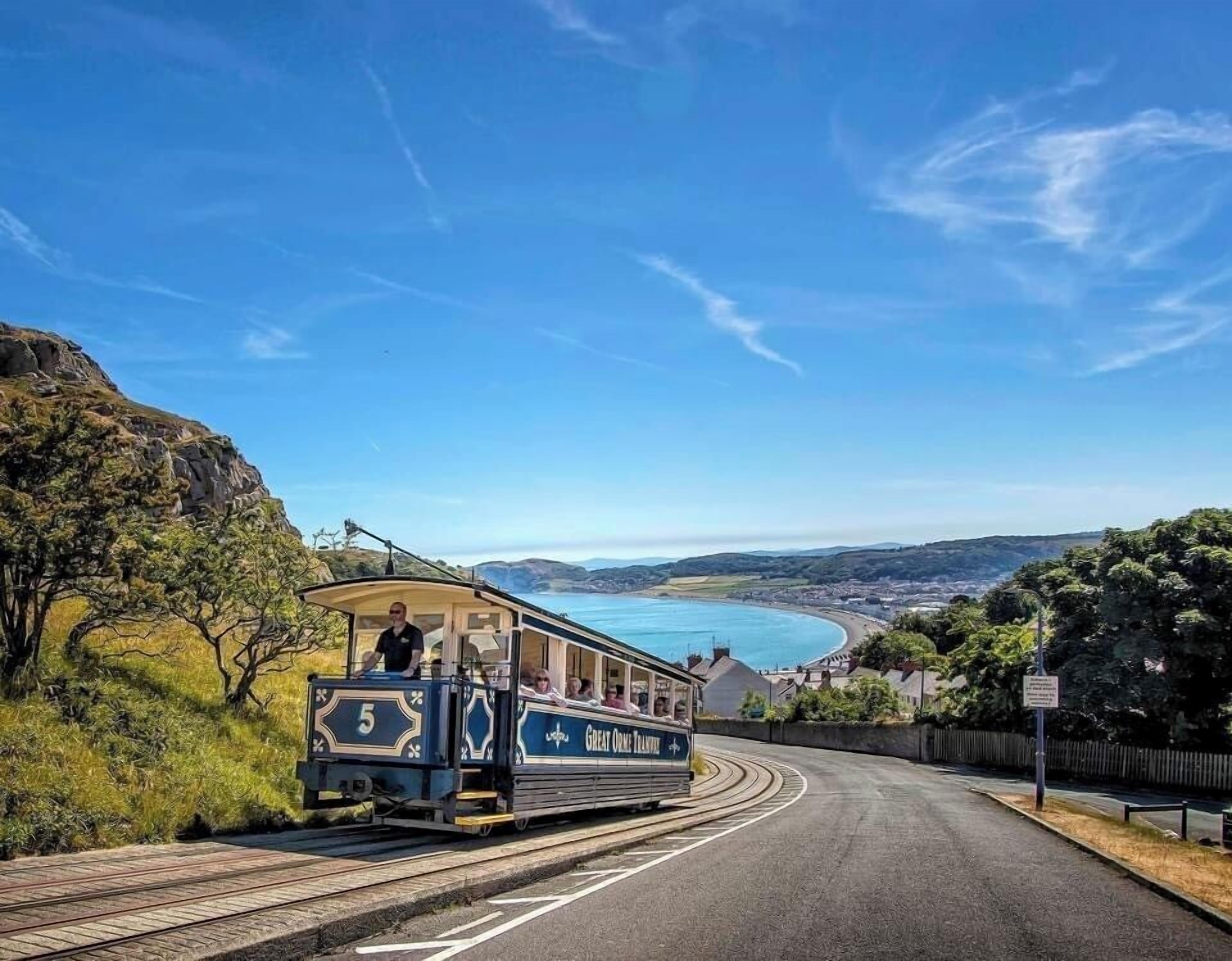 A tram with the number 5 and 'Great Orme Tram' written on it, traveling on tracks alongside a road with a coastal view.