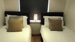 Two single beds with white linens and patterned pillows, a window with a nightlight, and a wooden floor.