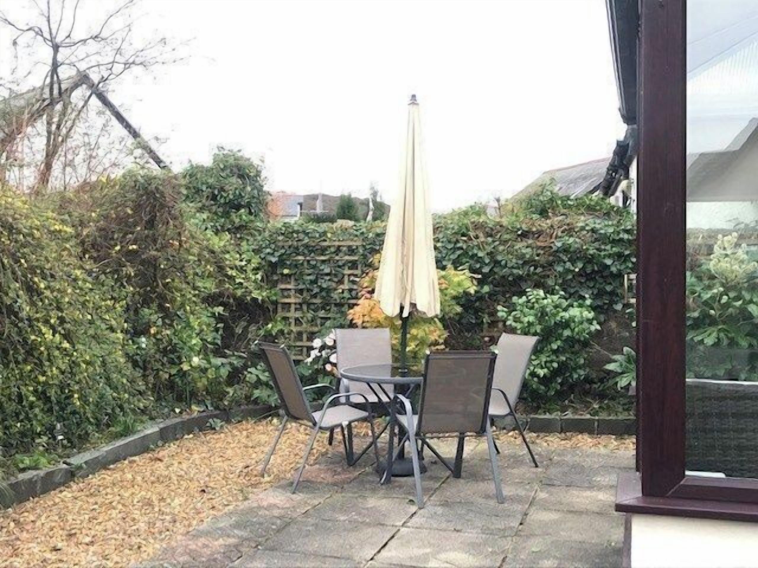 En udendørs patio med et bord og stole, omgivet af frodig beplantning og en hæk.