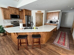 A kitchen with wooden cabinets, a central island with stools, stainless steel appliances, and a red patterned rug. - 1BR Teton View Getaway w/ Hot Tub near Skiing & Nat’l Parks (Driggs)
