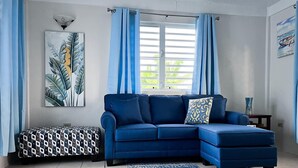 Una sala de estar con un sofá azul, un reposapiés estampado, una alfombra azul y un cuadro de hojas en la pared.