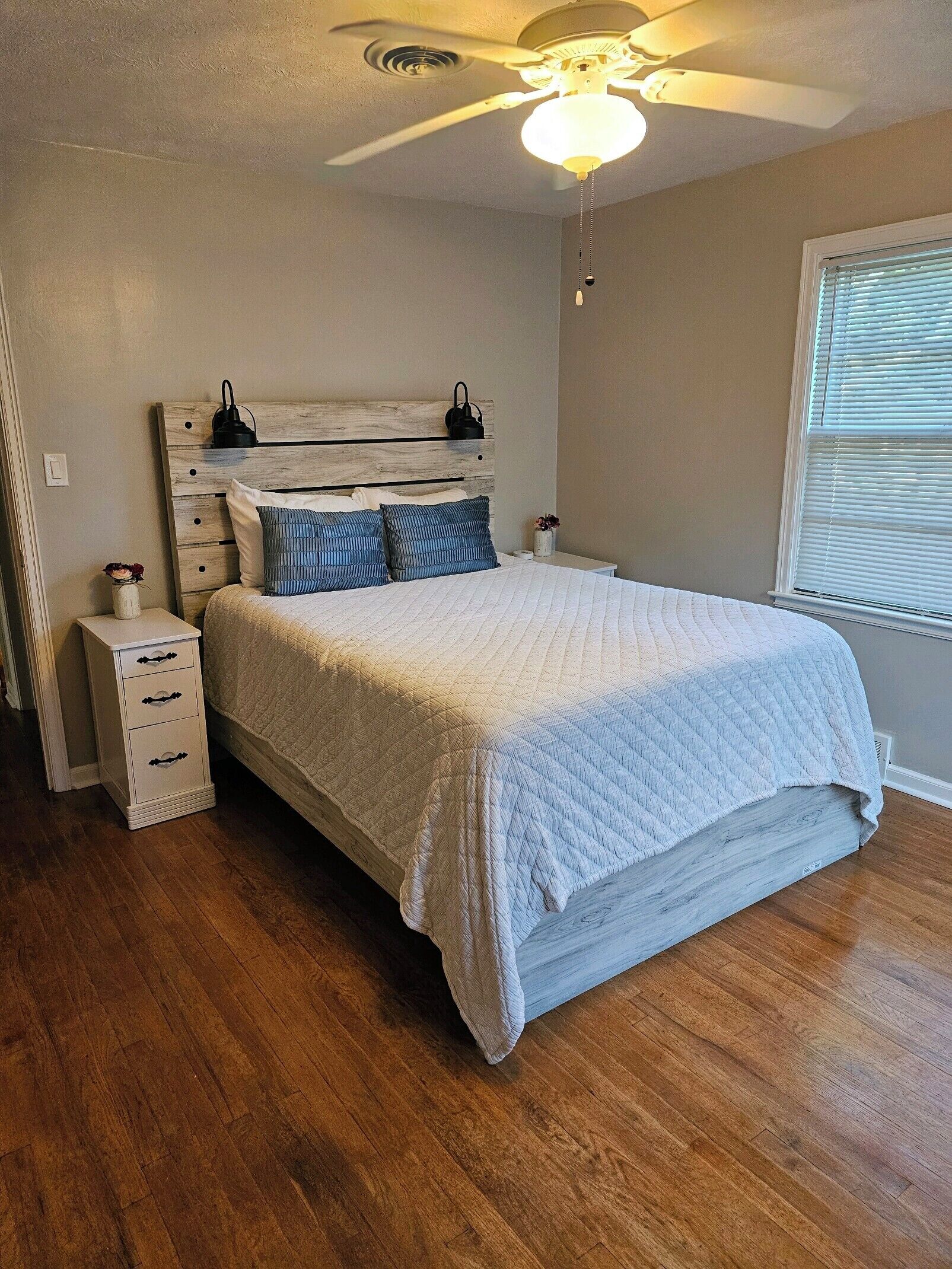 Een slaapkamer met een bed, nachtkastje, dressoir, plafondventilator en raam met jaloezieën.