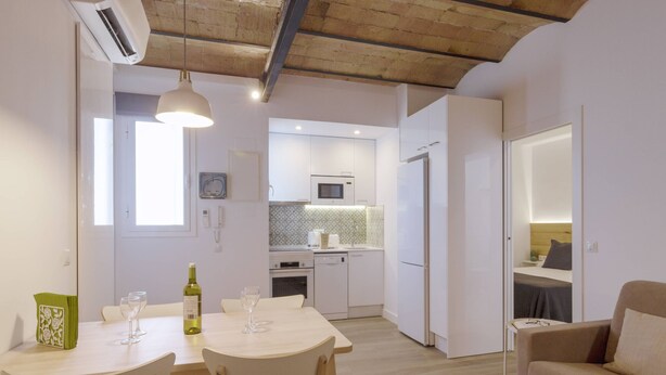 Una cocina moderna con armarios blancos, un microondas y un lavavajillas. Hay una mesa de comedor con sillas y una botella de vino.