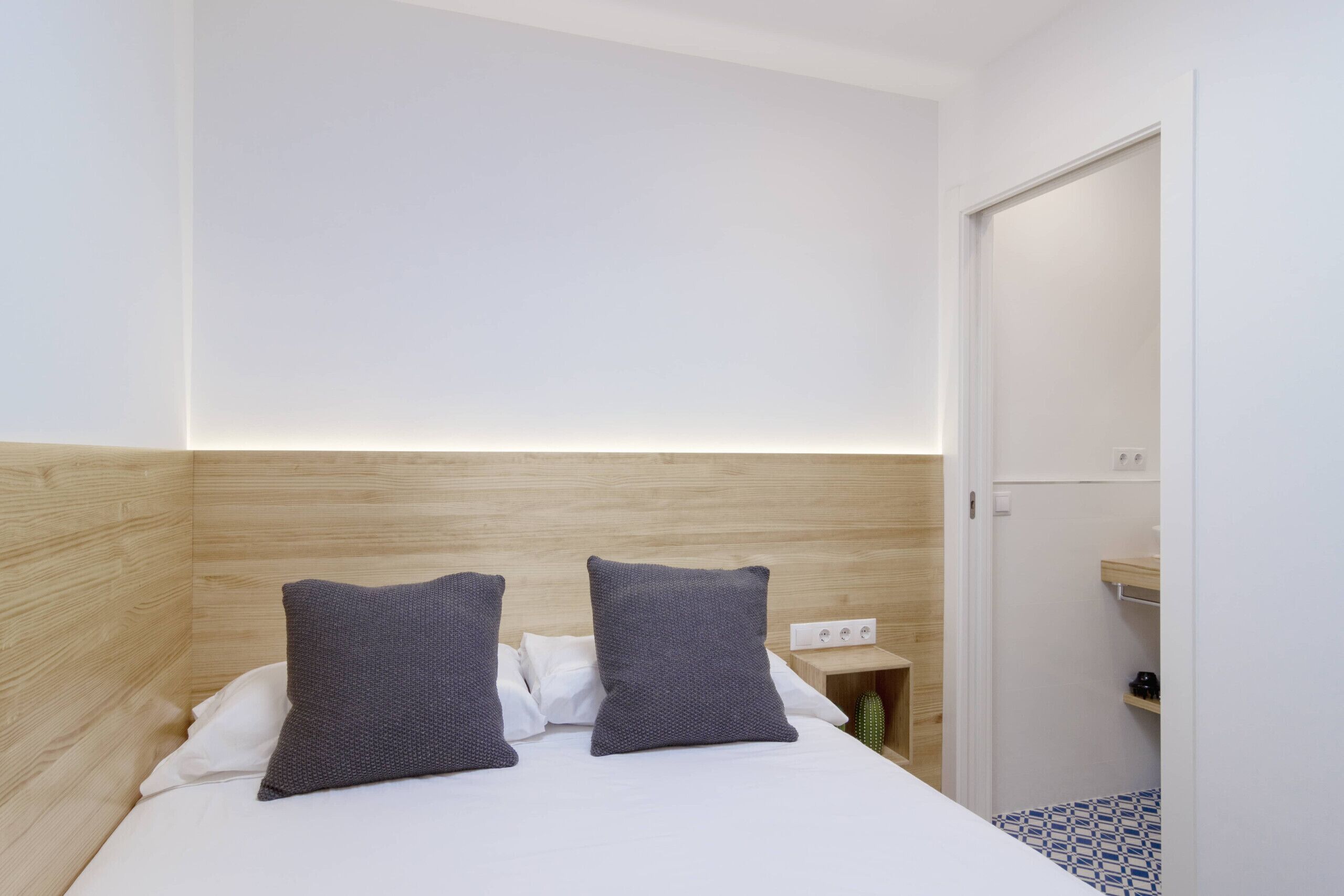 Habitación de hotel minimalista con cabecera de madera, dos almohadas grises, cubrecama blanco y una mesita de noche con lámpara.