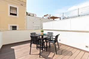 Terraza en la azotea con una mesa redonda y cuatro sillas, una planta pequeña en maceta, y una pared blanca con una ventana y una puerta.