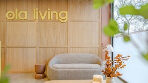 A cozy seating area with a sofa and a small table, a wooden wall with 'ola living' signage, and a window with a view of trees.