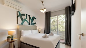A modern hotel room with a large bed, a bedside table with a lamp, a wall-mounted artwork, and a view of the outdoors through a large window.