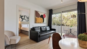 A modern living room with a black leather sofa, a glass coffee table, and a dining area with a white table and chairs. There is a large window providing a view of trees outside.