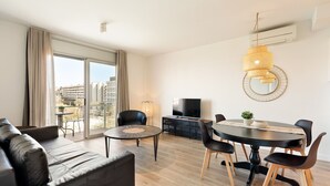 Ein modernes Wohnzimmer mit einer Couch, einem Esstisch mit Stühlen, einem Fernseher und einem großen Fenster mit Vorhängen.