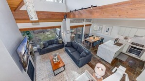 A spacious living area with a large sofa, a coffee table, and a dining table. There is a kitchen with a sink and stove. The room has wooden beams on the ceiling and large windows. - 3BD/3BA Village Townhome/Hot Tub (Whistler)