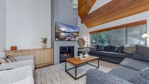 A modern living room with a fireplace, a sofa set, a coffee table, and a large window with a view of trees. - 3BD/3BA Village Townhome/Hot Tub (Whistler)