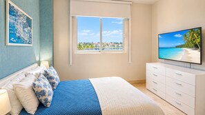 A bedroom with a bed, a television, a window with a view of a beach, and a painting on the wall.
