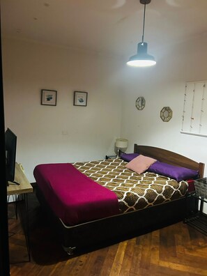 A bedroom with a bed, a desk, a chair, a TV, and a hanging light fixture.