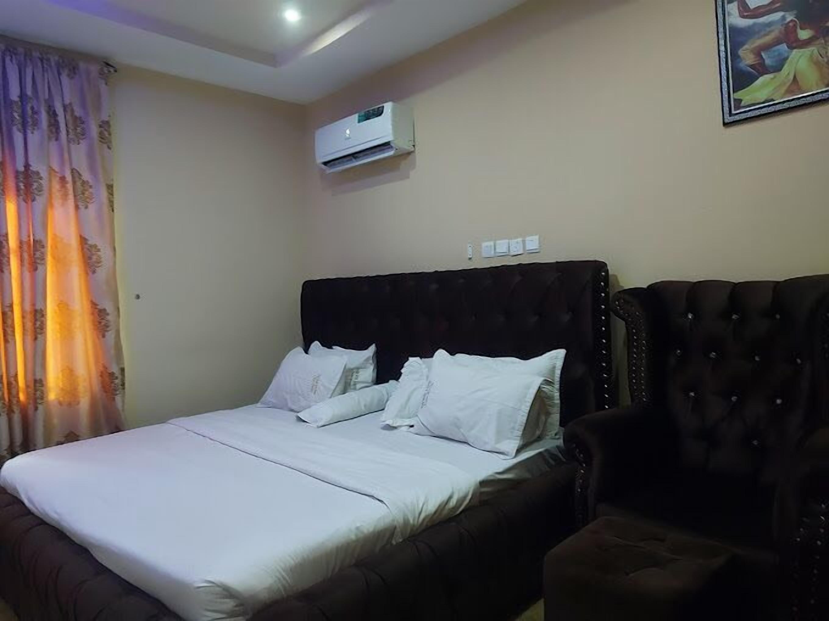 A bedroom with a bed, a dark upholstered armchair, an air conditioning unit, and a painting on the wall.