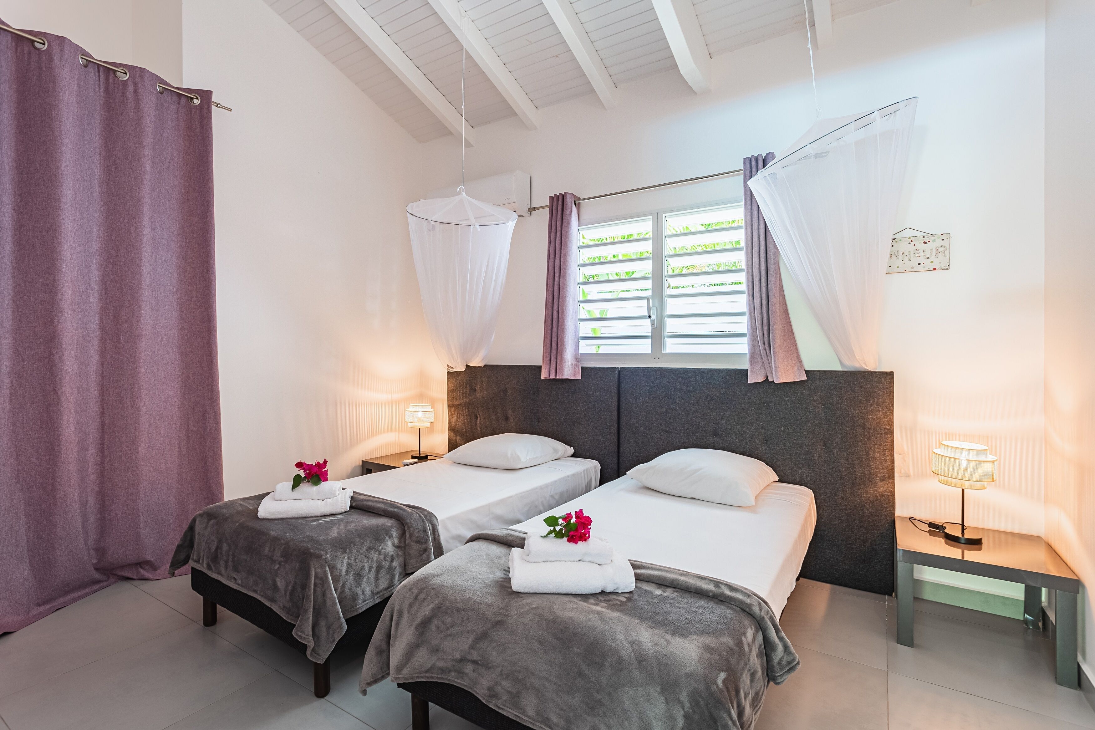 A hotel room with two beds, a window with curtains, a bedside table with a lamp, and a wall-mounted air conditioner.