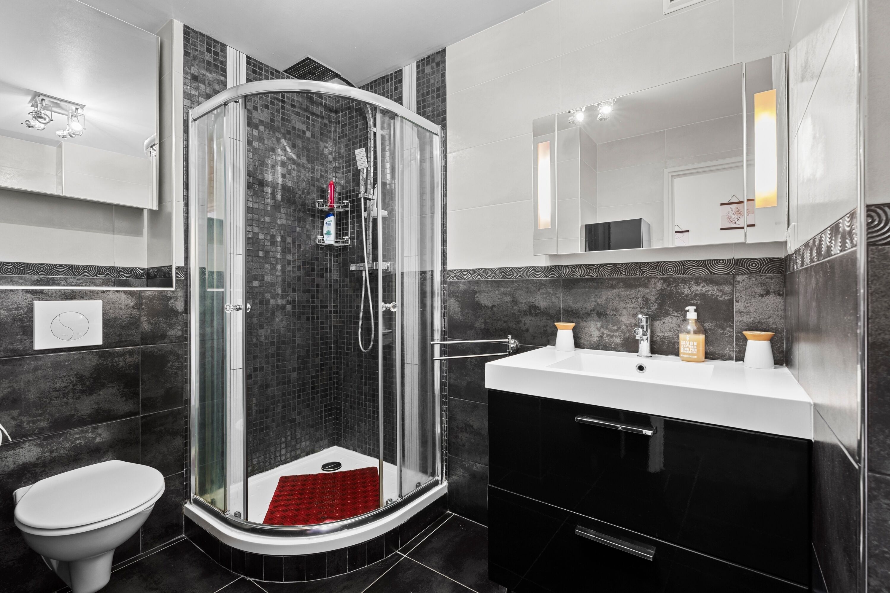 Un bany modern amb plat de dutxa envoltat de vidre, un lavabo blanc i mobles negres.