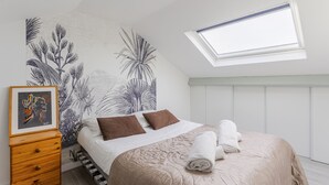 Une chambre à coucher avec un lit, une lucarne, un mur orné d’une fresque représentant des plantes, et une commode en bois sur laquelle est posée une photo encadrée.