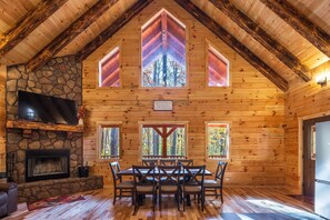A log cabin interior with a stone fireplace, wooden dining table, and large windows.