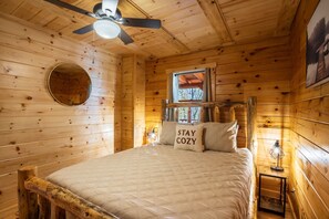A cozy wooden cabin room with a bed, a ceiling fan, a round mirror, a window with curtains, and a wall-mounted lamp.
