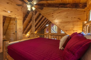 Ein Schlafzimmer in einer Holzhütte mit roter Bettdecke, Holzbalken, Deckenventilator und einem Fenster mit Aussicht.