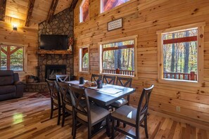 Interior de una cabaña de madera con chimenea de piedra, una mesa de comedor con sillas y grandes ventanales con vistas al bosque.
