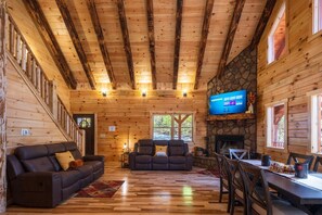 Interior acogedor de una cabaña con paredes de madera, una chimenea de piedra, un televisor montado en la pared y una zona de comedor con mesa y sillas.