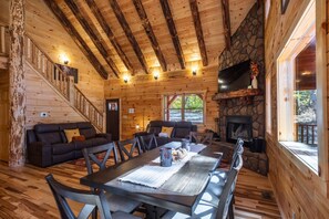 Interior de una cabaña de troncos con chimenea de piedra, paredes de madera, un comedor y una escalera.