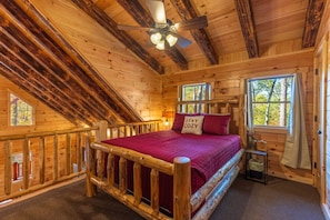 Un dormitorio rústico tipo cabaña de madera, con una cama de madera, ventilador de techo y ventana con cortinas.