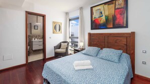 A bedroom with a wooden headboard, a bed with a blue patterned bedspread, a white towel, a chair, a mirror, and a large abstract painting on the wall. - Villa Lanzabur - Three Bedroom Villa, Sleeps 6 (Teguise)