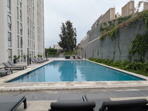 A rectangular swimming pool with lounge chairs and a tall tree in the background.