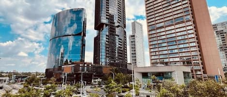 Zona urbana moderna con edificios altos, una estructura de vidrio curva y un paseo peatonal con jardines amarillos.