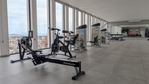Un gimnasio con diverso equipo de ejercicio, amplias ventanas y vista a un paisaje urbano.