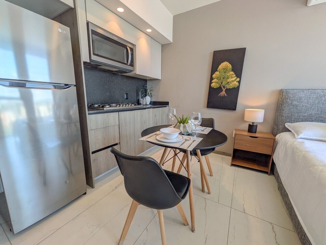 A modern hotel room with a kitchenette, a dining area with two chairs, a bed, and a nightstand with a lamp.