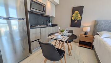 A modern hotel room with a kitchenette, a dining area with two chairs, a bed, and a nightstand with a lamp.