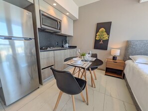 Een moderne hotelkamer met een kitchenette, een eethoek met twee stoelen, een bed en een nachtkastje met een lamp.